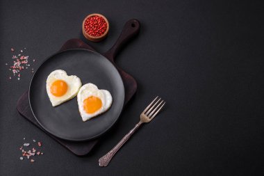 Two heart-shaped fried eggs on a black ceramic plate on a dark concrete background. Breakfast for valentine's day