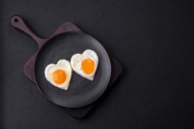 Two heart-shaped fried eggs on a black ceramic plate on a dark concrete background. Breakfast for valentine's day