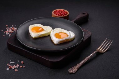Two heart-shaped fried eggs on a black ceramic plate on a dark concrete background. Breakfast for valentine's day