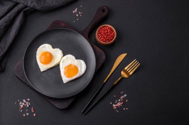 Two heart-shaped fried eggs on a black ceramic plate on a dark concrete background. Breakfast for valentine's day