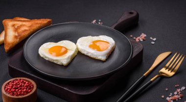 Two heart-shaped fried eggs on a black ceramic plate on a dark concrete background. Breakfast for valentine's day