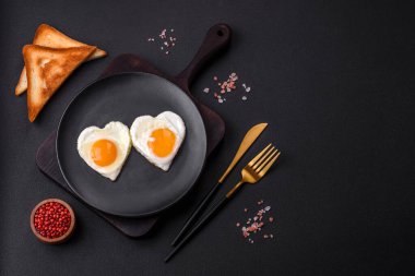 Two heart-shaped fried eggs on a black ceramic plate on a dark concrete background. Breakfast for valentine's day