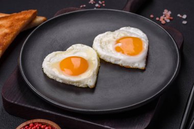 Two heart-shaped fried eggs on a black ceramic plate on a dark concrete background. Breakfast for valentine's day