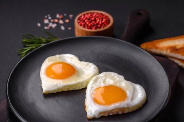 Two heart-shaped fried eggs on a black ceramic plate on a dark concrete background. Breakfast for valentine's day