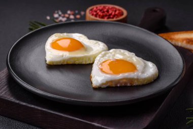 Two heart-shaped fried eggs on a black ceramic plate on a dark concrete background. Breakfast for valentine's day