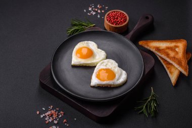 Two heart-shaped fried eggs on a black ceramic plate on a dark concrete background. Breakfast for valentine's day