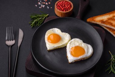 Two heart-shaped fried eggs on a black ceramic plate on a dark concrete background. Breakfast for valentine's day