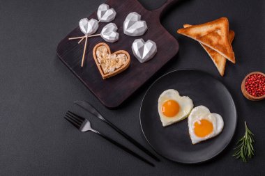 Two heart-shaped fried eggs on a black ceramic plate on a dark concrete background. Breakfast for valentine's day