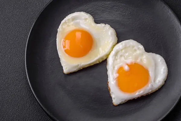 Two heart-shaped fried eggs on a black ceramic plate on a dark concrete background. Breakfast for valentine's day