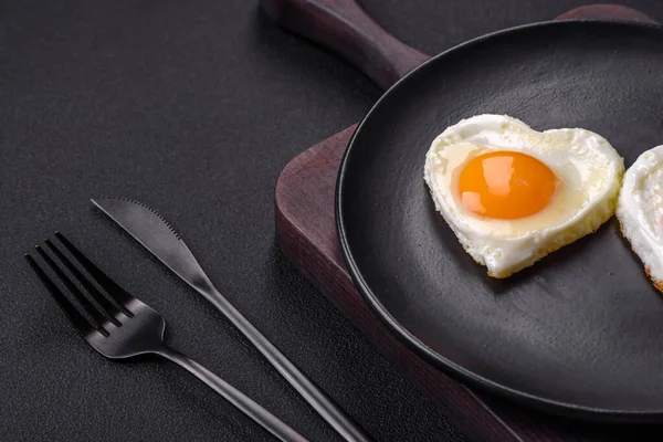 Two heart-shaped fried eggs on a black ceramic plate on a dark concrete background. Breakfast for valentine's day