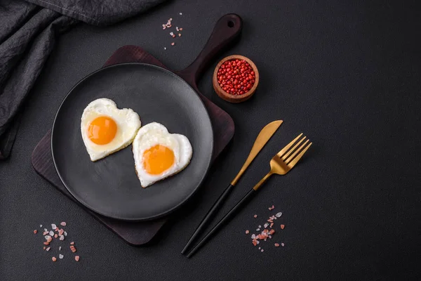 Two heart-shaped fried eggs on a black ceramic plate on a dark concrete background. Breakfast for valentine's day