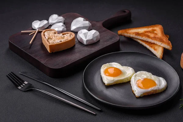 Two heart-shaped fried eggs on a black ceramic plate on a dark concrete background. Breakfast for valentine's day