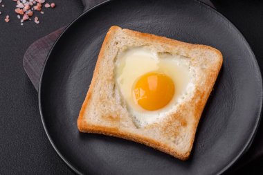 Kalp şeklinde kızarmış yumurta ve susamlı tost, keten tohumu ve balkabağı çekirdeği siyah bir tabakta koyu beton arka planda.