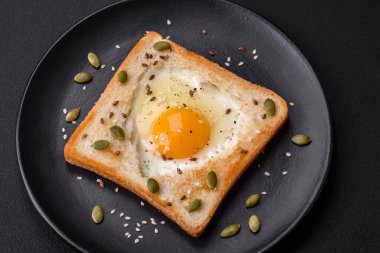 Kalp şeklinde kızarmış yumurta ve susamlı tost, keten tohumu ve balkabağı çekirdeği siyah bir tabakta koyu beton arka planda.