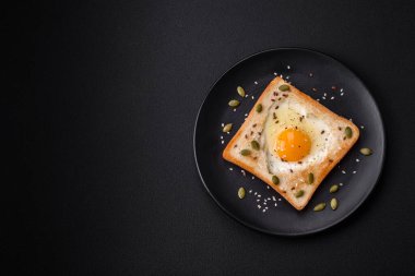 Kalp şeklinde kızarmış yumurta ve susamlı tost, keten tohumu ve balkabağı çekirdeği siyah bir tabakta koyu beton arka planda.