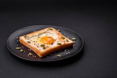 Kalp şeklinde kızarmış yumurta ve susamlı tost, keten tohumu ve balkabağı çekirdeği siyah bir tabakta koyu beton arka planda.