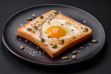 Kalp şeklinde kızarmış yumurta ve susamlı tost, keten tohumu ve balkabağı çekirdeği siyah bir tabakta koyu beton arka planda.