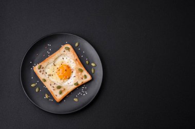 Kalp şeklinde kızarmış yumurta ve susamlı tost, keten tohumu ve balkabağı çekirdeği siyah bir tabakta koyu beton arka planda.