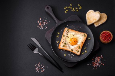 Kalp şeklinde kızarmış yumurta ve susamlı tost, keten tohumu ve balkabağı çekirdeği siyah bir tabakta koyu beton arka planda.
