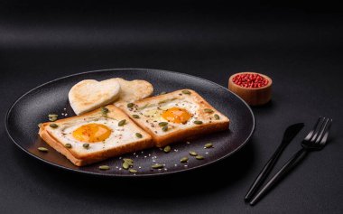 Kalp şeklinde kızarmış yumurta ve susamlı tost, keten tohumu ve balkabağı çekirdeği siyah bir tabakta koyu beton arka planda.