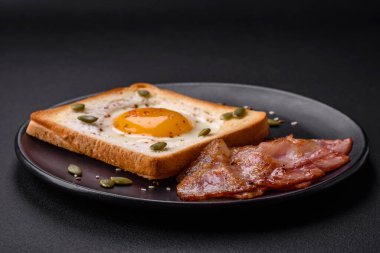 Kalp şeklinde kızarmış yumurta ve susamlı tost, keten tohumu ve balkabağı çekirdeği siyah bir tabakta koyu beton arka planda.