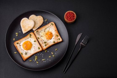 Kalp şeklinde kızarmış yumurta ve susamlı tost, keten tohumu ve balkabağı çekirdeği siyah bir tabakta koyu beton arka planda.