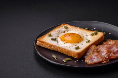 Kalp şeklinde kızarmış yumurta ve susamlı tost, keten tohumu ve balkabağı çekirdeği siyah bir tabakta koyu beton arka planda.