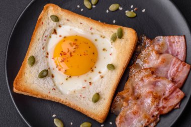 Kalp şeklinde kızarmış yumurta ve susamlı tost, keten tohumu ve balkabağı çekirdeği siyah bir tabakta koyu beton arka planda.