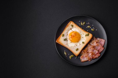 Kalp şeklinde kızarmış yumurta ve susamlı tost, keten tohumu ve balkabağı çekirdeği siyah bir tabakta koyu beton arka planda.