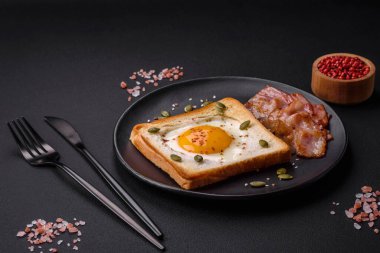 Kalp şeklinde kızarmış yumurta ve susamlı tost, keten tohumu ve balkabağı çekirdeği siyah bir tabakta koyu beton arka planda.