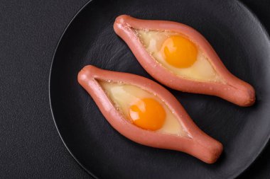 Composite concept of fried eggs inside a cut sausage with spices on a black ceramic plate on a dark concrete background