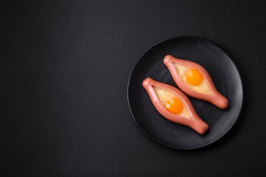 Composite concept of fried eggs inside a cut sausage with spices on a black ceramic plate on a dark concrete background