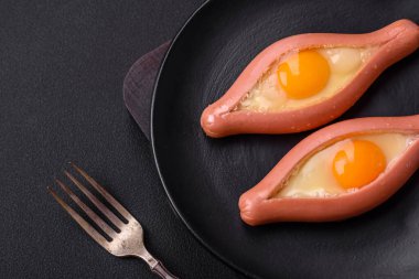 Composite concept of fried eggs inside a cut sausage with spices on a black ceramic plate on a dark concrete background