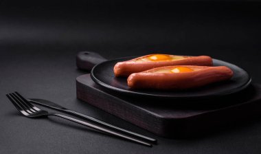 Composite concept of fried eggs inside a cut sausage with spices on a black ceramic plate on a dark concrete background