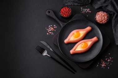 Composite concept of fried eggs inside a cut sausage with spices on a black ceramic plate on a dark concrete background