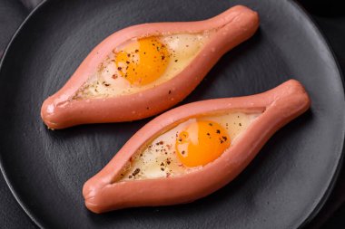 Composite concept of fried eggs inside a cut sausage with spices on a black ceramic plate on a dark concrete background