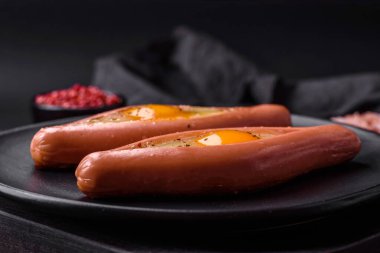 Composite concept of fried eggs inside a cut sausage with spices on a black ceramic plate on a dark concrete background