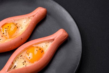 Composite concept of fried eggs inside a cut sausage with spices on a black ceramic plate on a dark concrete background
