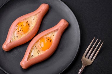 Composite concept of fried eggs inside a cut sausage with spices on a black ceramic plate on a dark concrete background
