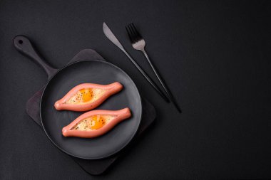Composite concept of fried eggs inside a cut sausage with spices on a black ceramic plate on a dark concrete background
