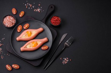 Composite concept of fried eggs inside a cut sausage with spices on a black ceramic plate on a dark concrete background