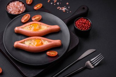 Composite concept of fried eggs inside a cut sausage with spices on a black ceramic plate on a dark concrete background