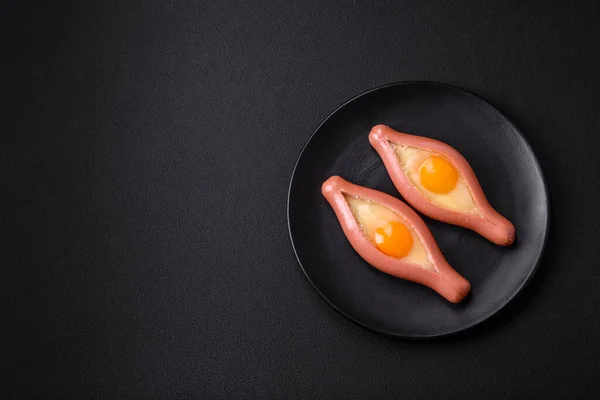 Composite concept of fried eggs inside a cut sausage with spices on a black ceramic plate on a dark concrete background