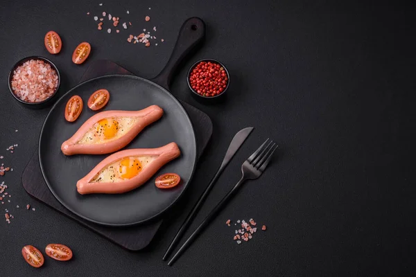 Composite concept of fried eggs inside a cut sausage with spices on a black ceramic plate on a dark concrete background