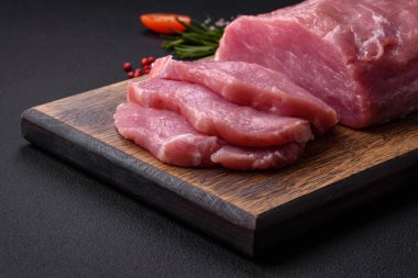 A piece of raw fresh pork on a wooden cutting board with spices and herbs on a dark concrete background
