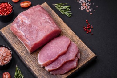 A piece of raw fresh pork on a wooden cutting board with spices and herbs on a dark concrete background