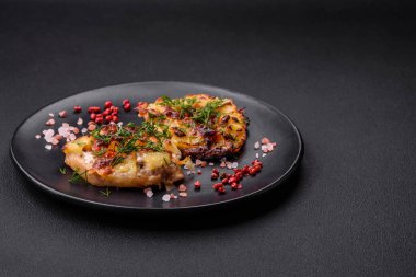 Meat baked in the oven with pineapple, tomatoes, cheese, spices and herbs on a black ceramic plate on a dark concrete background