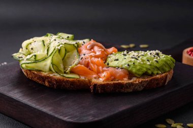 Delicious brown bread toast with salmon, avocado, cucumber and sesame seeds on a textured concrete background