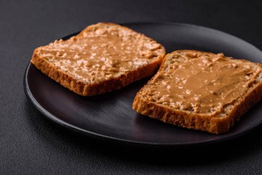 Nutritious sandwich consisting of bread and peanut butter on a black ceramic plate on a dark concrete background