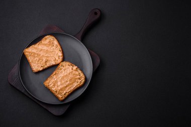 Nutritious sandwich consisting of bread and peanut butter on a black ceramic plate on a dark concrete background